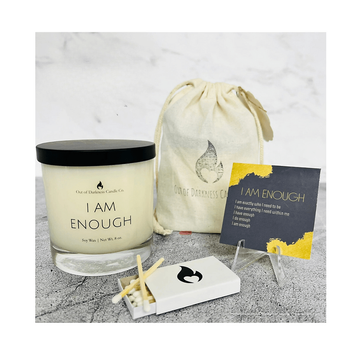 Affirmation candle set by Out of Darkness Candle Co. featuring a candle labeled 'I AM ENOUGH', a matchbox, and a card with affirmations. The set includes a drawstring bag with the company's logo and a gray, marbled background.