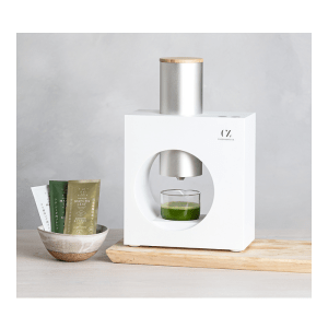 Modern and sleek matcha tea maker with a white body and a cylindrical silver top, placed on a wooden surface. The front of the machine has a circular cutout where a glass cup filled with green matcha tea is positioned. Next to the matcha maker, there is a ceramic bowl containing three different colored matcha tea packets: one green, one gold, and one white. The background is a smooth gray wall, adding to the minimalist aesthetic.