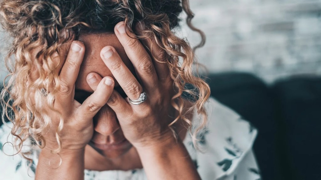 Woman with curly hair holding her head in frustration, representing the emotional distress of parental estrangement discussed in the article "Why Don’t You Speak to Your Parents?" The article explores the societal taboo of severing parental bonds, the judgment faced by estranged individuals, and the broader emotional costs and family impacts of estrangement.