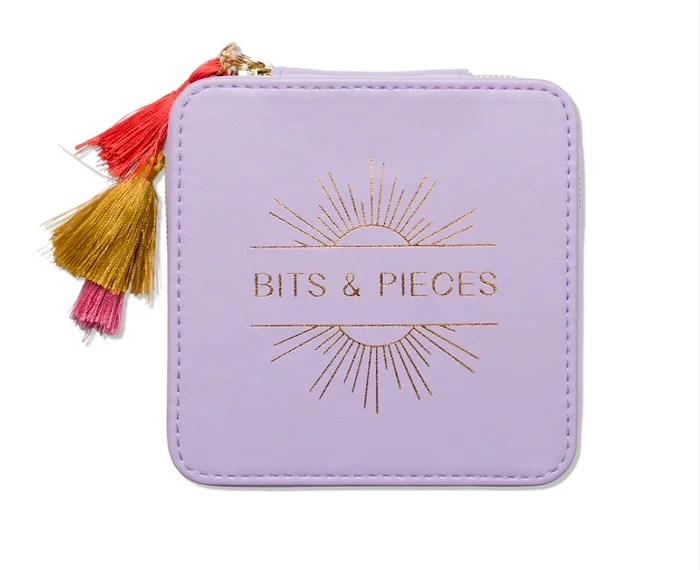 Lavender-colored jewelry box with 'BITS & PIECES' written in gold, accented by a sunburst design. The box has tassels in red, gold, and pink attached to the zipper, adding a playful touch.