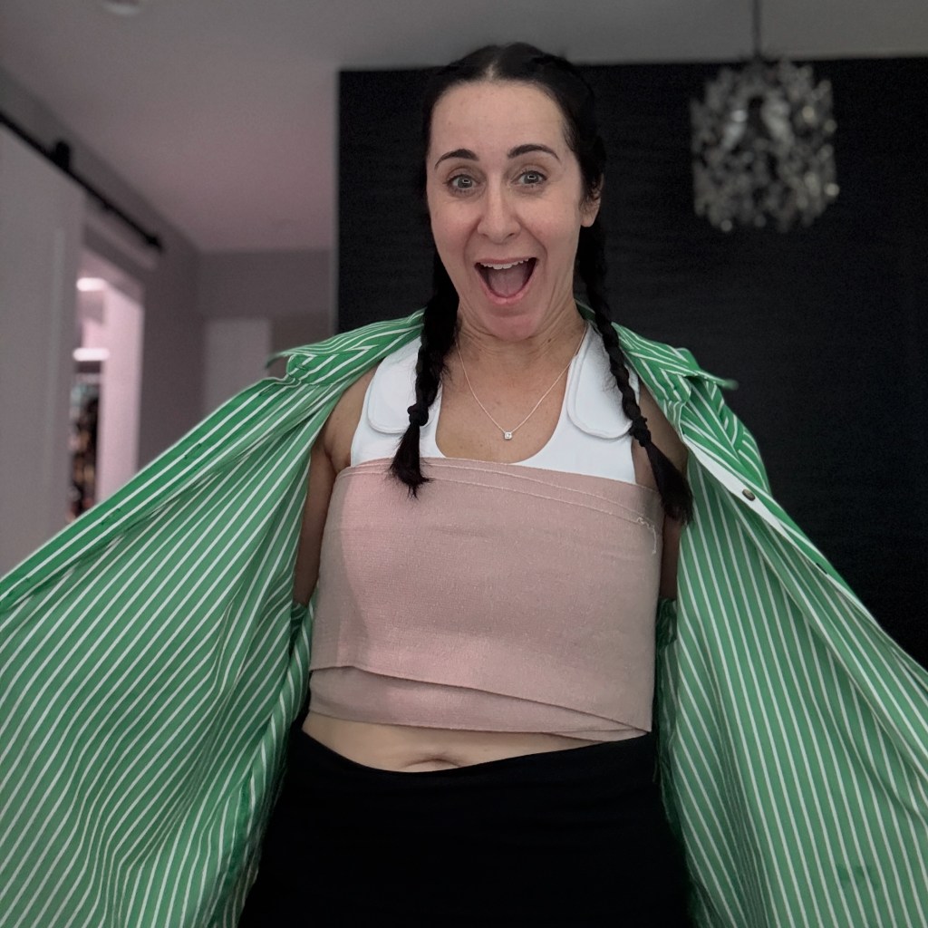 A woman with braided hair smiles and opens her green striped shirt, revealing a soft pink wrap around her torso. The background is neutral, enhancing her expression of joy and confidence post-surgery.