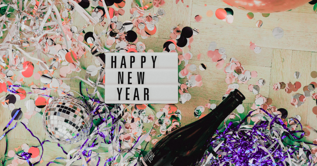 Overhead view of a New Year’s celebration scene with colorful confetti, metallic streamers, a small disco ball, an empty champagne bottle, and a lightbox sign that reads ‘Happy New Year’ scattered across a wooden floor.