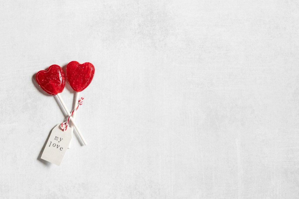Two red heart shaped lollipops with a white tag that says "my love" on a light grey background symbolizing two people in a marriage on Valentine's Day