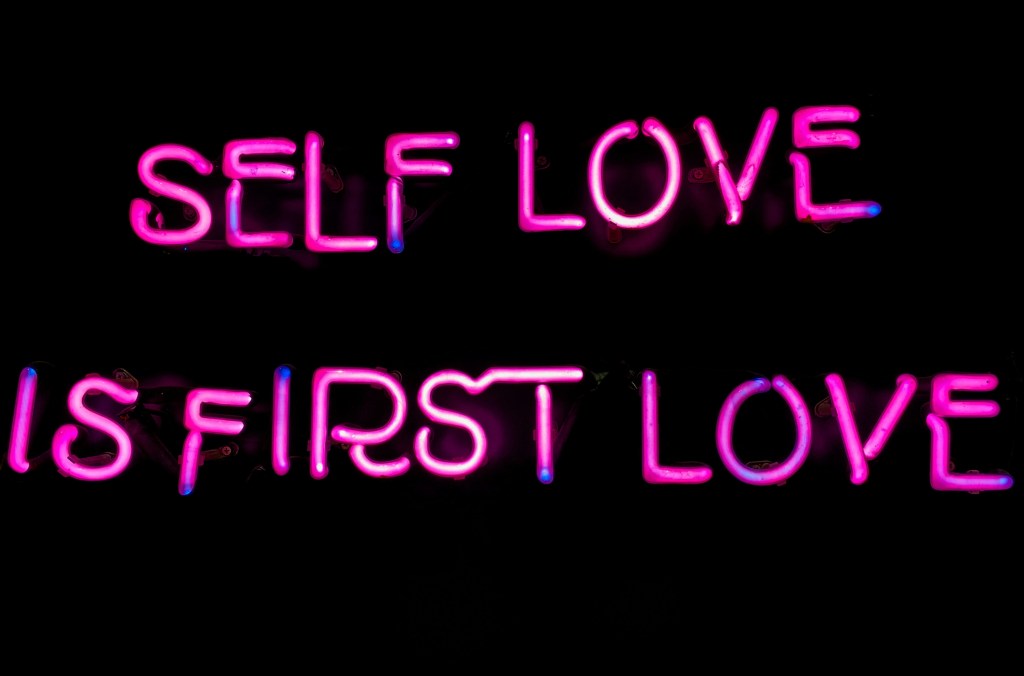 A neon sign that says self love is first love in pink.