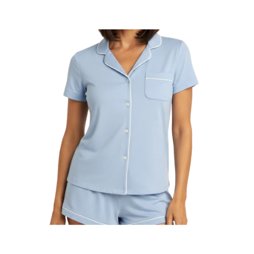 A woman wearing a light blue short-sleeve pajama set with a button-up top and matching shorts, featuring a pocket and white piping from Lusomé.