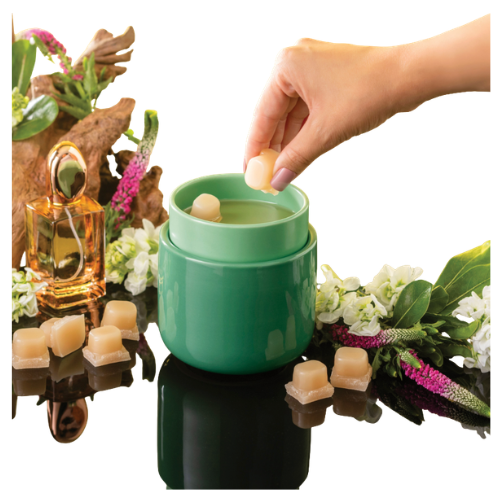 A hand reaching into a green container to pick up wax melts, surrounded by decorative flowers and a bottle of fragrance.
