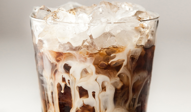 A glass filled with iced coffee, featuring ice cubes and swirls of cream mixing with the dark beverage.