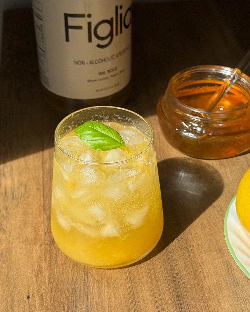 A refreshing glass of non-alcoholic beverage with ice, garnished with a basil leaf, placed on a wooden surface next to a jar of honey and a whole lemon.