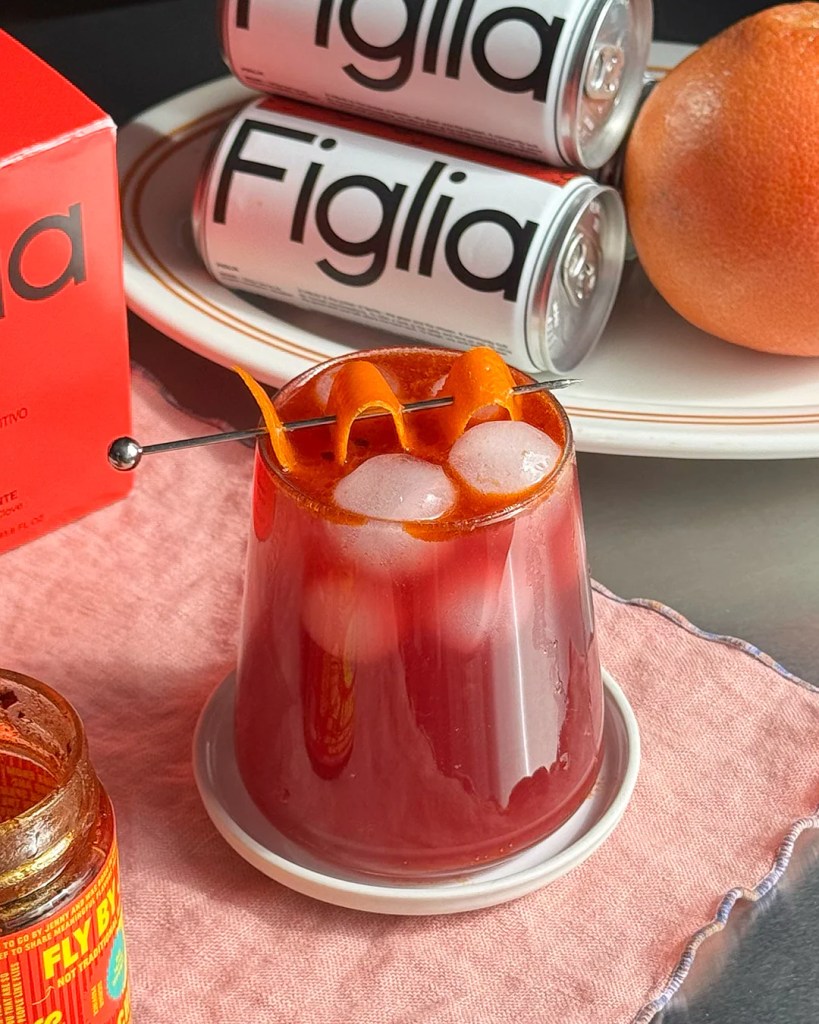 A vibrant cocktail in a glass with ice, garnished with orange twists, served on a small white plate. In the background, there are cans of Figlia and a grapefruit.