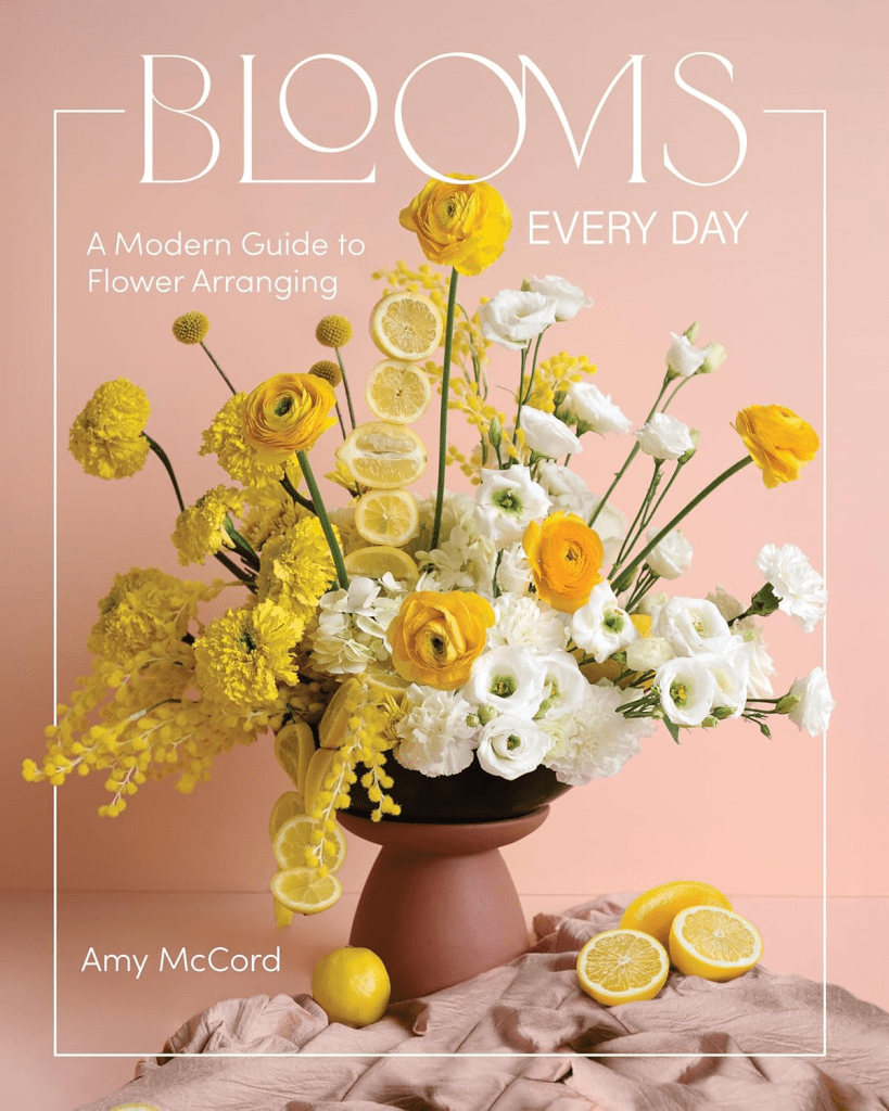 A vibrant flower arrangement featuring yellow and white flowers, including ranunculus and lisianthus, complemented by fresh lemons, displayed in a terracotta pot against a soft pink background.