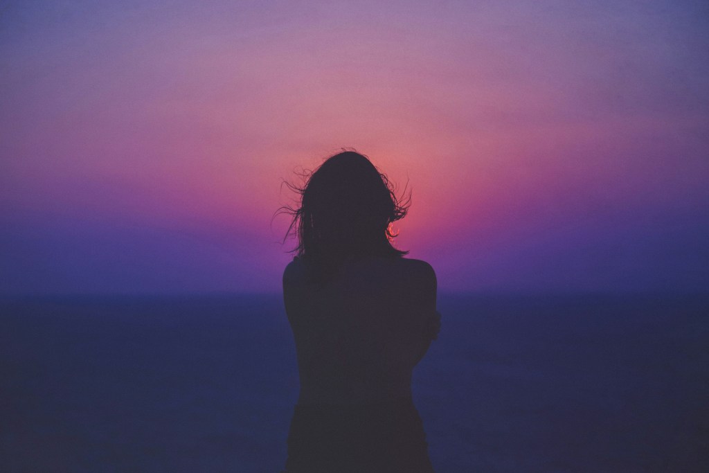 Silhouette of a person standing by the ocean during a colorful sunset with hues of purple and pink.
