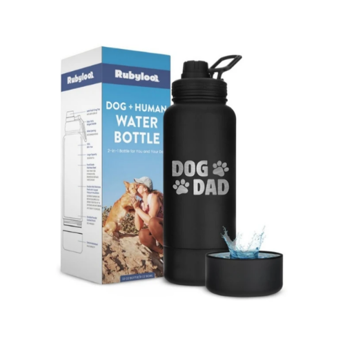 A black water bottle designed for both dogs and humans, with the text 'DOG DAD' printed on it. The bottle is placed next to its packaging, which features images of a dog and a human. A small water dish accompanies the bottle.