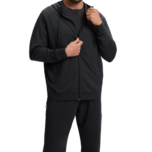 A man wearing a black zip-up hoodie and matching pants, with one hand on the zipper and the other resting at his side.