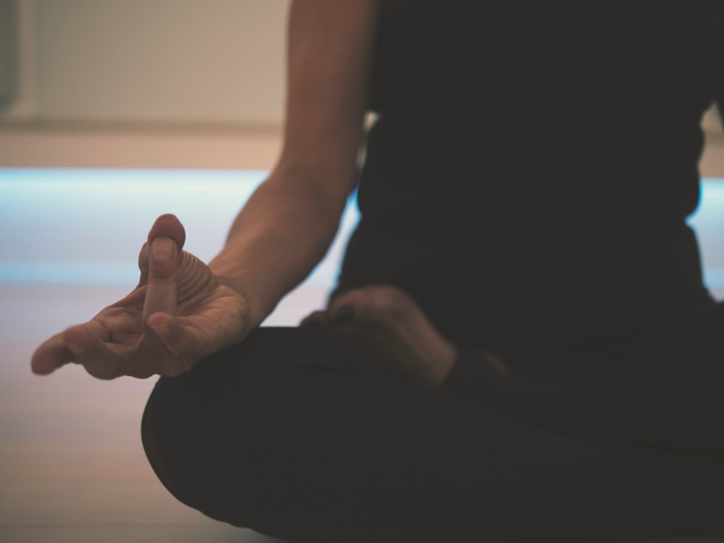 A close-up of a person's hand performing a meditation gesture, with the hand resting near the knee in a seated position, showcasing a calm and focused atmosphere.