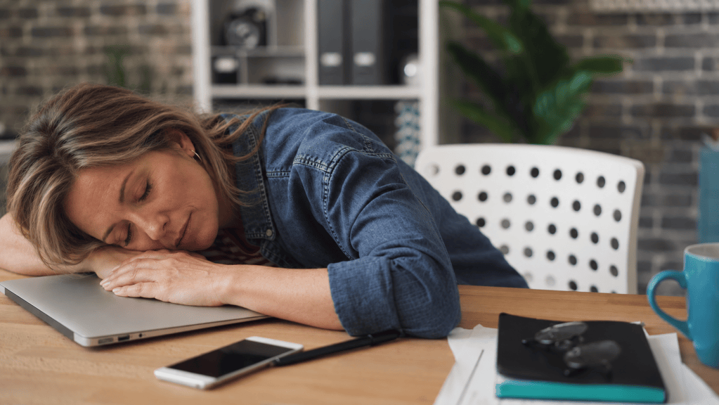 A woman resting her head on a laptop at a wooden desk, surrounded by a smartphone, a notebook, and a blue coffee mug.