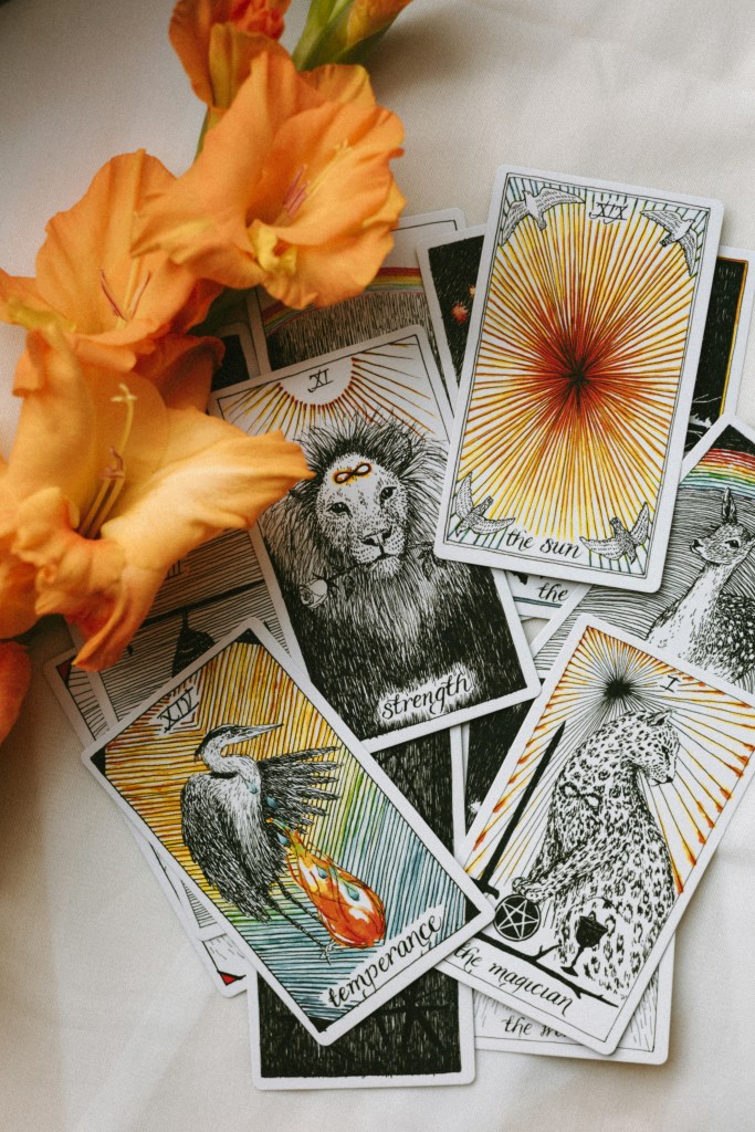 A collection of tarot cards displayed with vibrant illustrations, including 'Strength', 'Temperance', 'The Magician', and 'The Sun', alongside orange flowers on a light surface.